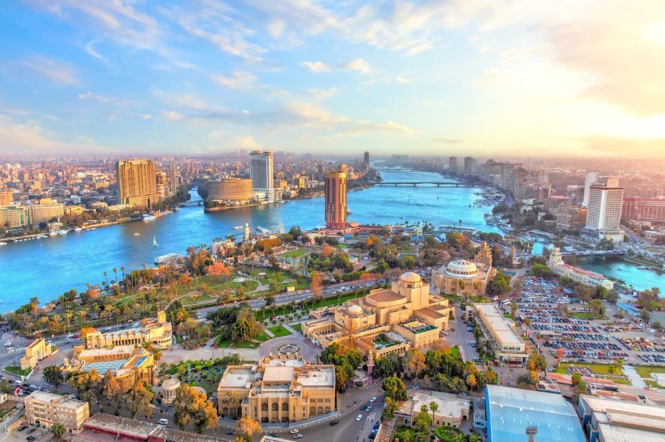 Vista aerea di una vasta metropoli attraversata da un ampio fiume blu, affiancata da edifici moderni e parchi verdi, sotto un cielo velato.