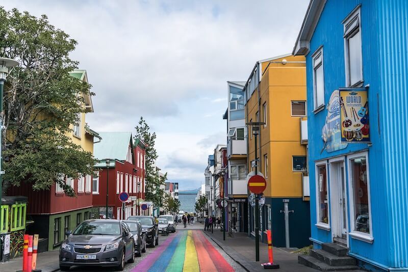 Una strada dipinta con un arcobaleno conduce verso il mare, fiancheggiata da edifici colorati e auto parcheggiate sotto un cielo nuvoloso.