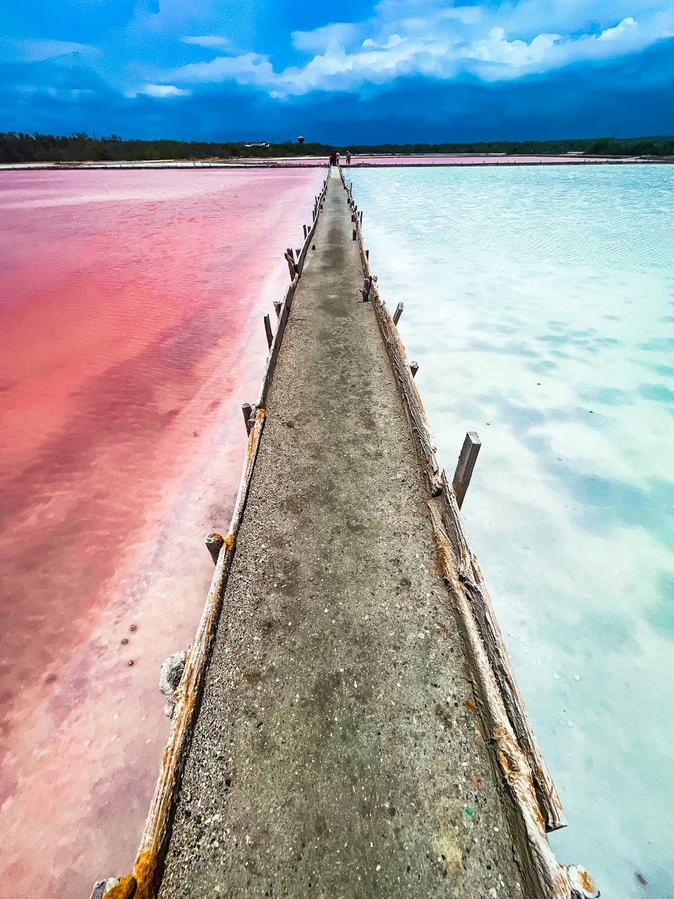 A long concrete pier divides a body of water, with vibrant pink water on one side and pale turquoise water on the other, under a blue sky.