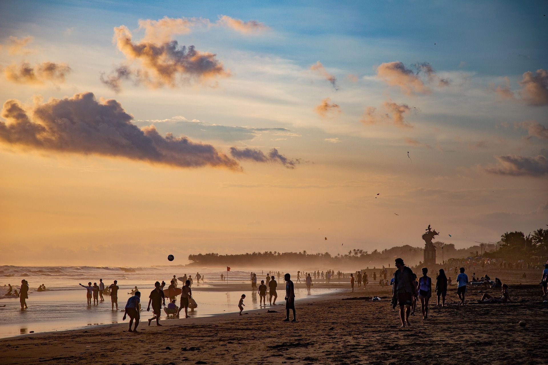 Sagome di persone su una spiaggia affollata al tramonto, che giocano e passeggiano in riva al mare sotto un cielo dorato.