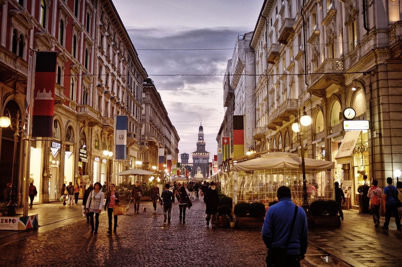 People walk down a cobblestone street lined with illuminated shops and flags at dusk, with a historic castle visible in the distance.