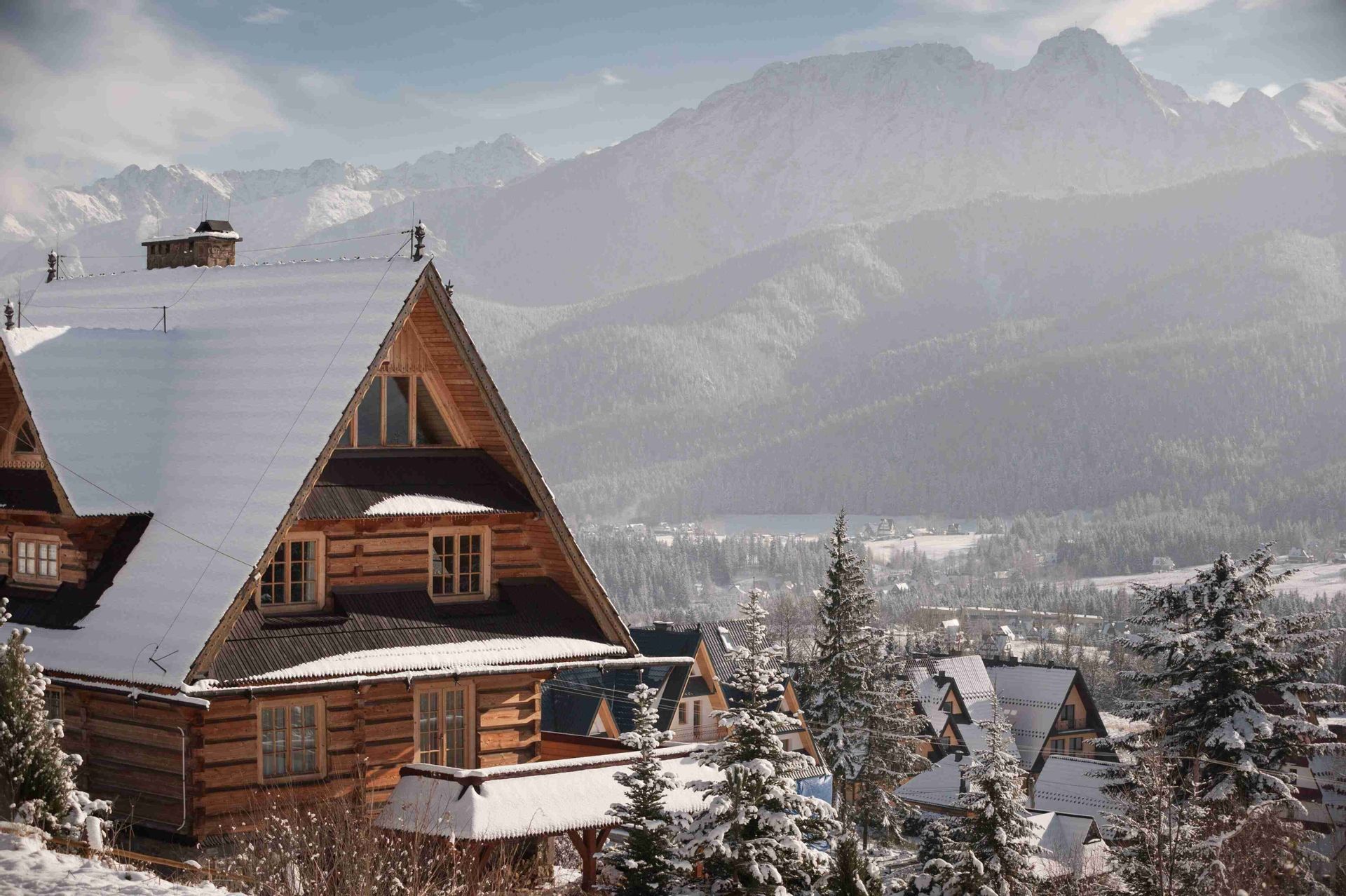Un chalet de madera con tejado nevado domina un pueblo de montaña y pinos, con grandes picos nevados de fondo.