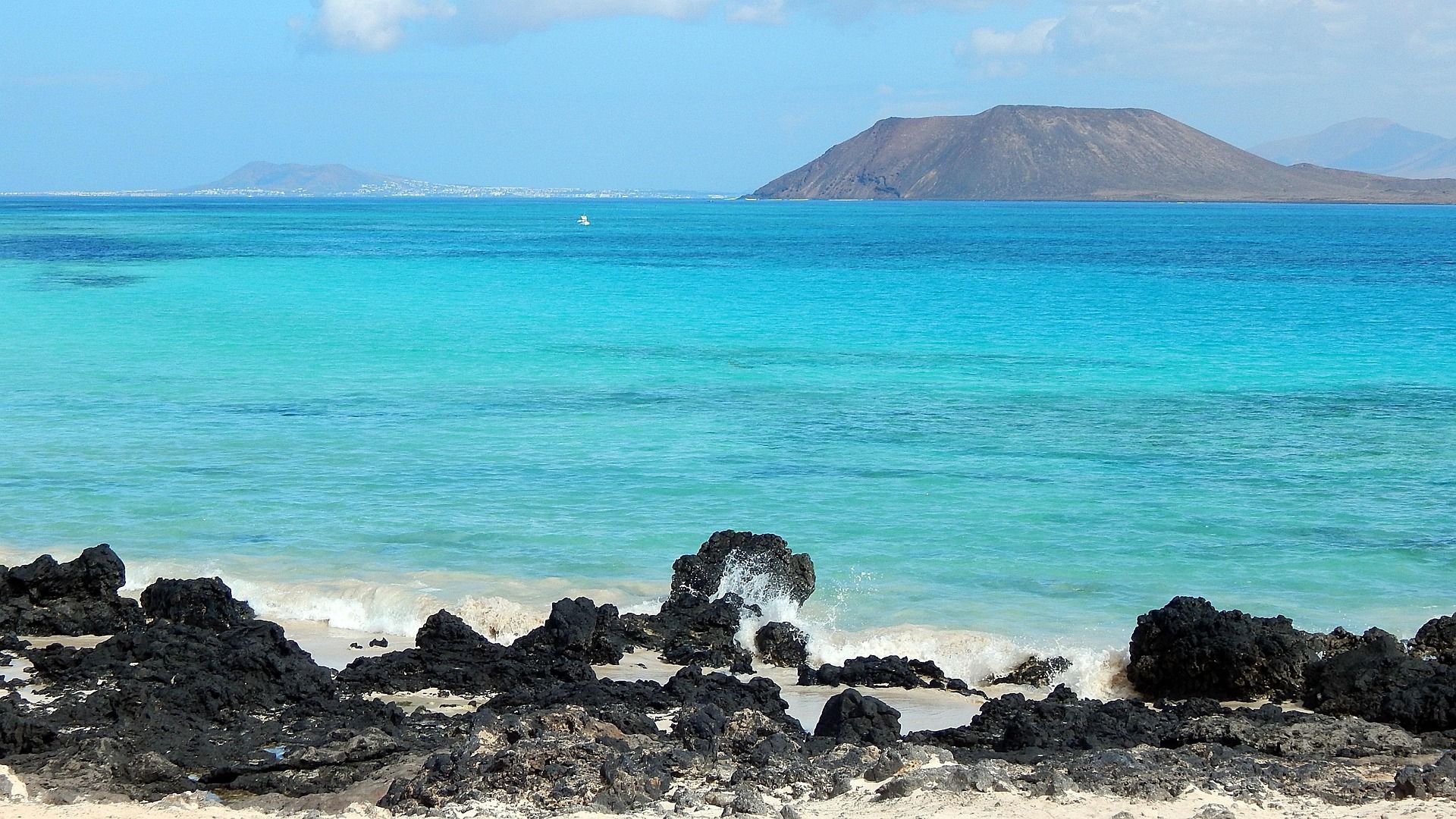 Las olas rompen contra rocas volcánicas negras en una playa de arena, con agua turquesa y una gran isla al fondo.