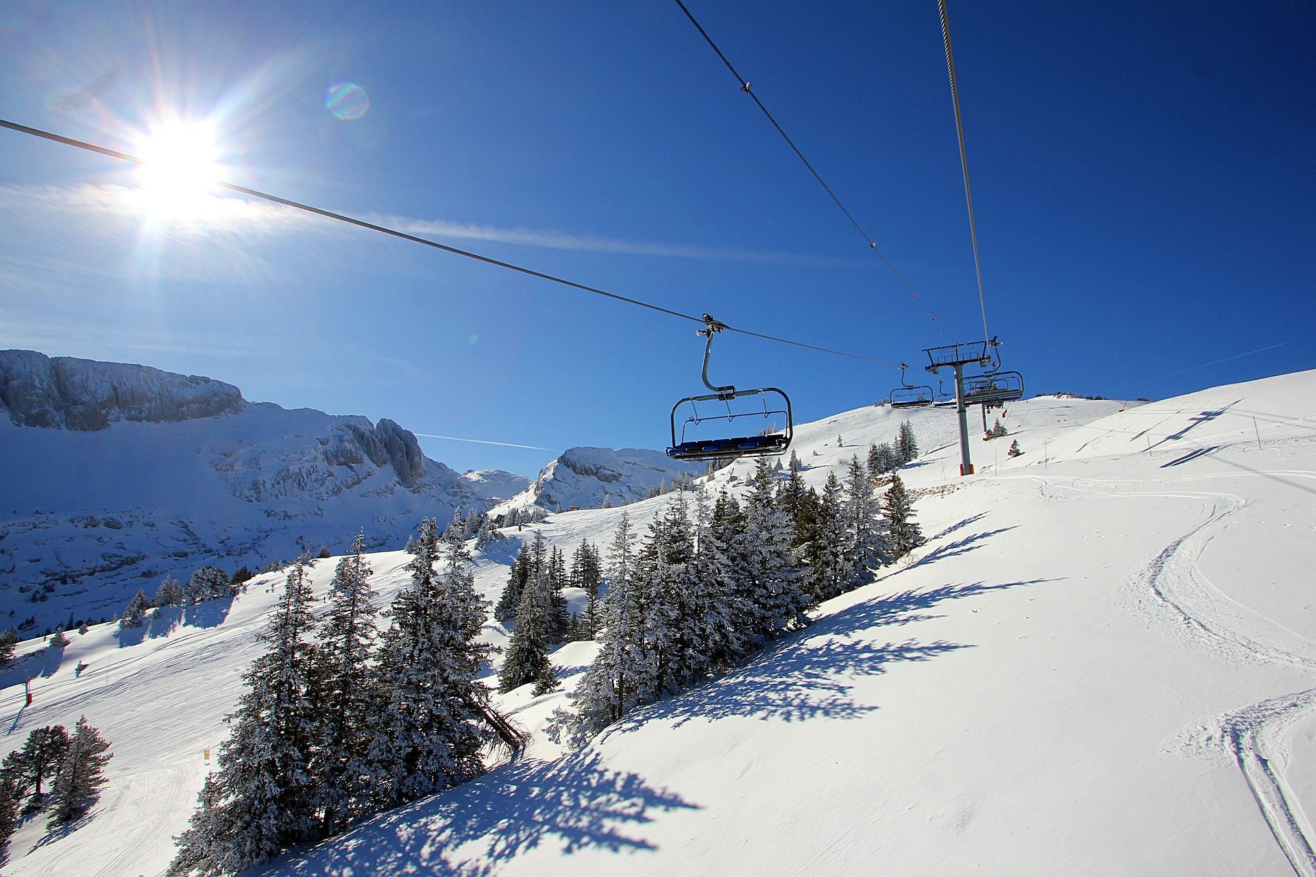 Un telesilla vacío asciende una ladera nevada con pinos abajo, bajo un sol brillante y un cielo azul claro.