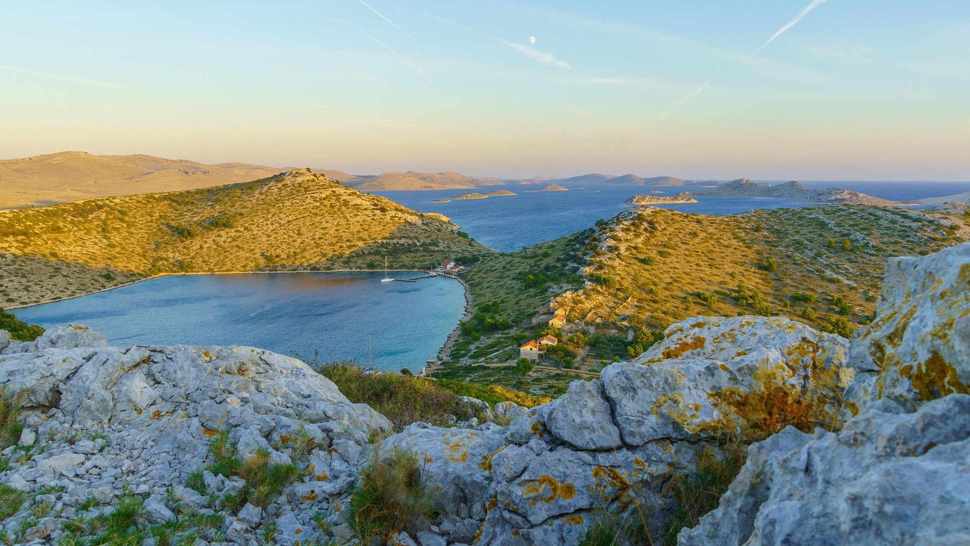 Una vista mozzafiato da una collina rocciosa che domina una tranquilla baia blu, circondata da colline e isole illuminate dal sole sotto un cielo limpido.