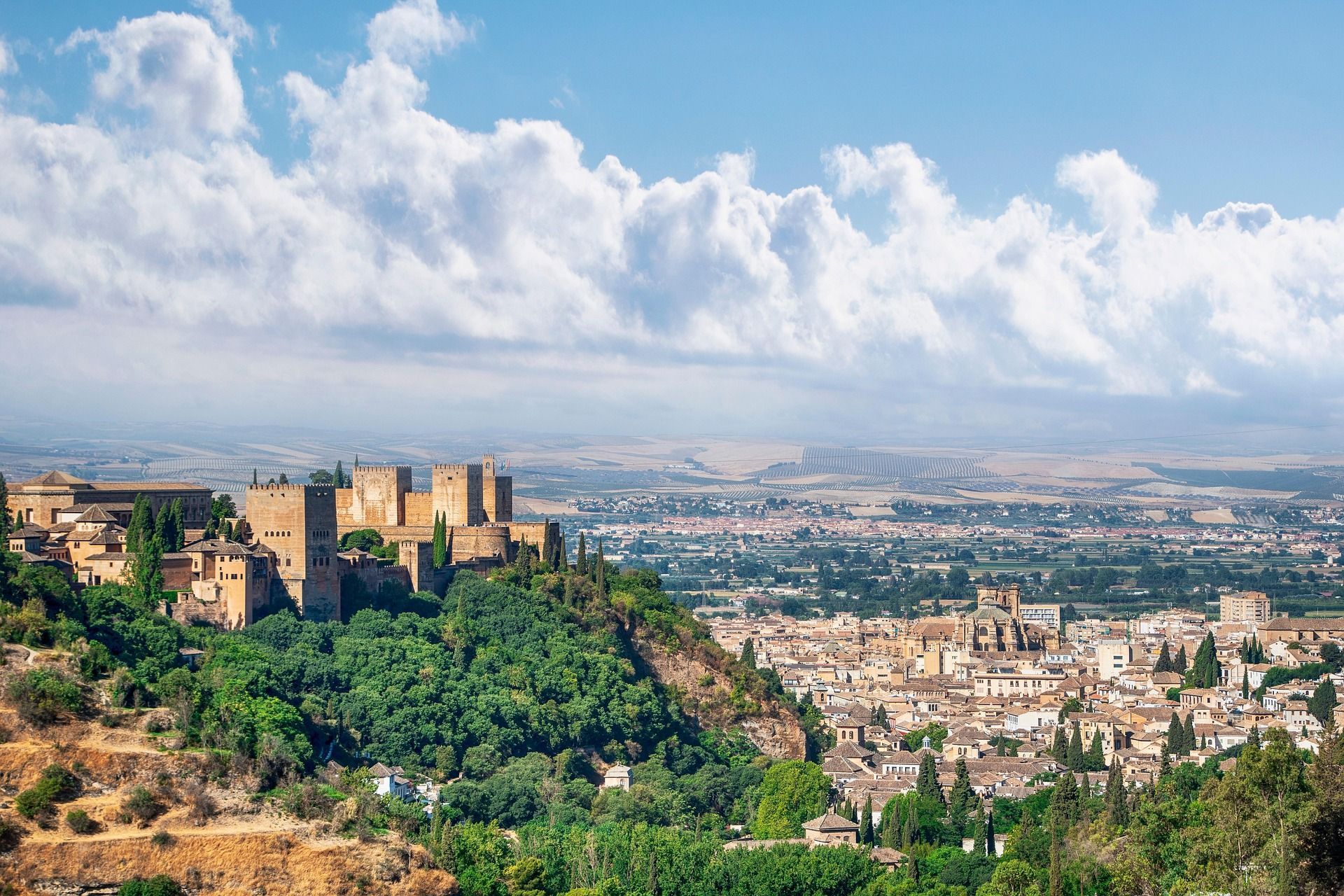 Una antigua fortaleza de piedra se asienta sobre una exuberante colina verde, dominando una extensa ciudad bajo un cielo azul con nubes esponjosas.