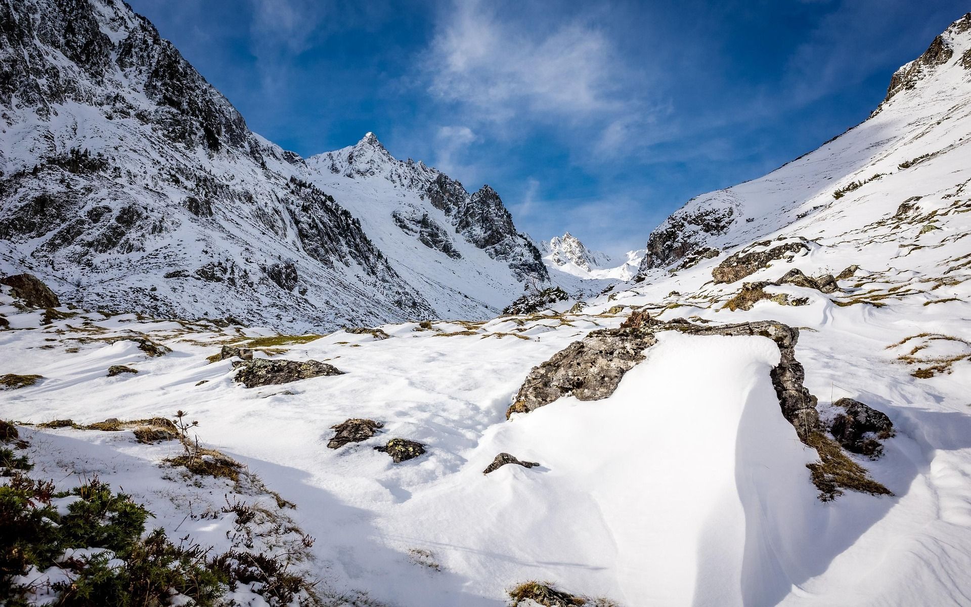 Un amplio valle montañoso cubierto de nieve con grandes rocas y picos escarpados bajo un cielo azul brillante con nubes tenues.