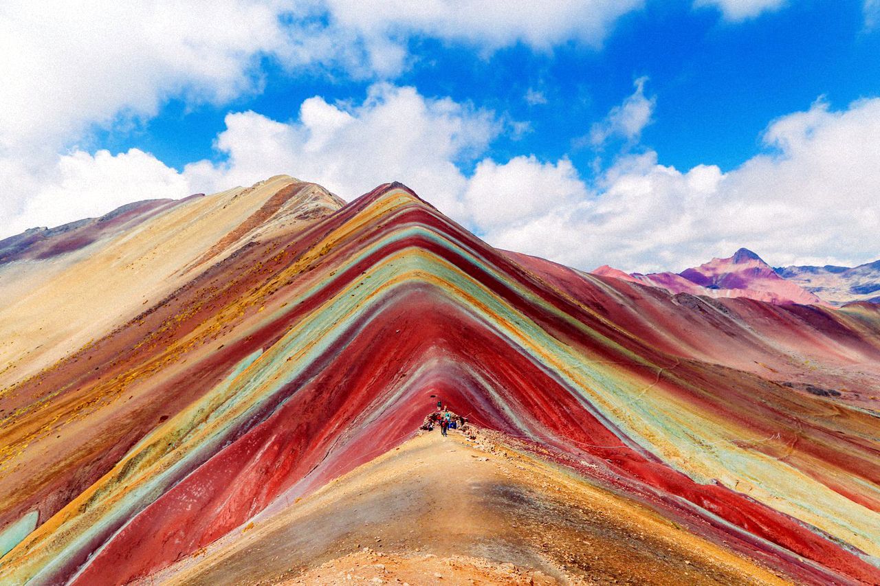 Un viaggio di gruppo WeRoad si trova sulla cresta di una grande montagna a strisce multicolori sotto un cielo azzurro con nuvole.