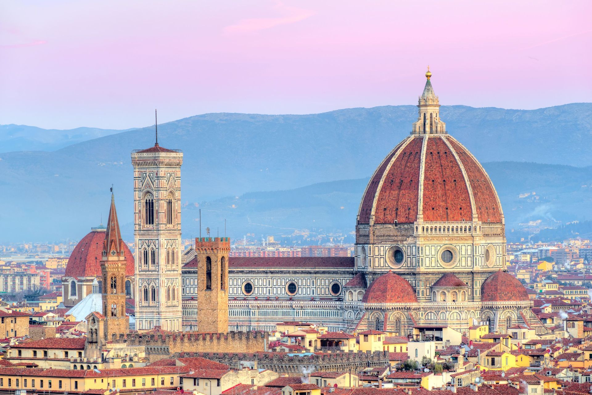 A large cathedral with a red-tiled dome and a tall bell tower overlooking a cityscape at dusk, with mountains in the distance.