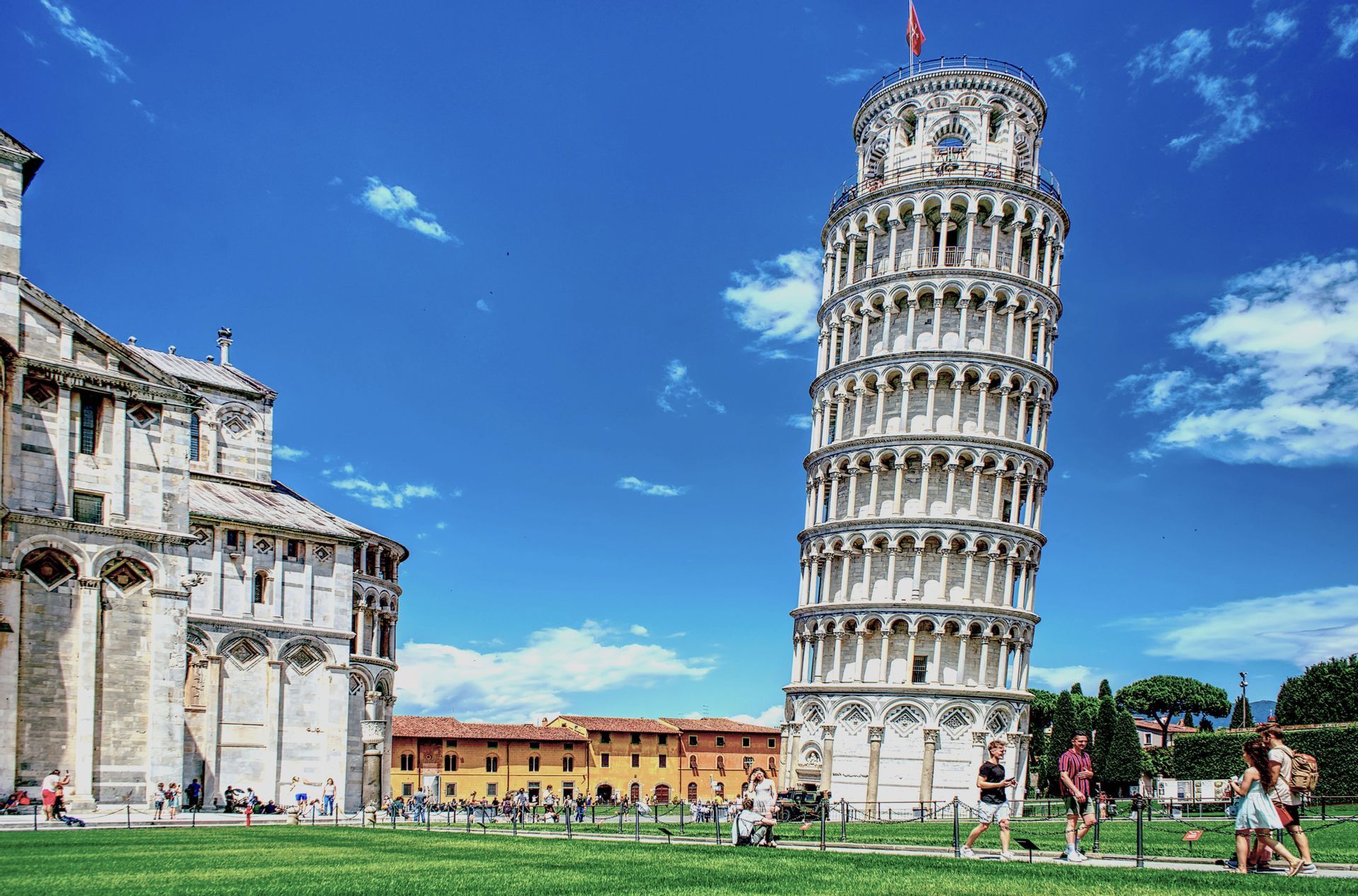 The Leaning Tower of Pisa and a cathedral stand on a green lawn with tourists walking by under a bright blue sky.
