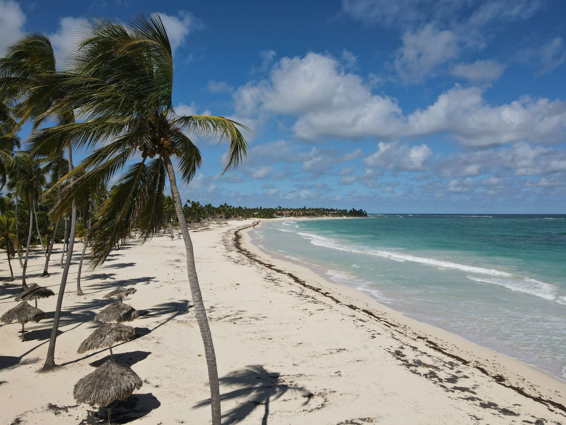 An aerial view of a white sand beach lined with palm trees and thatched umbrellas next to a turquoise ocean under a partly cloudy sky.