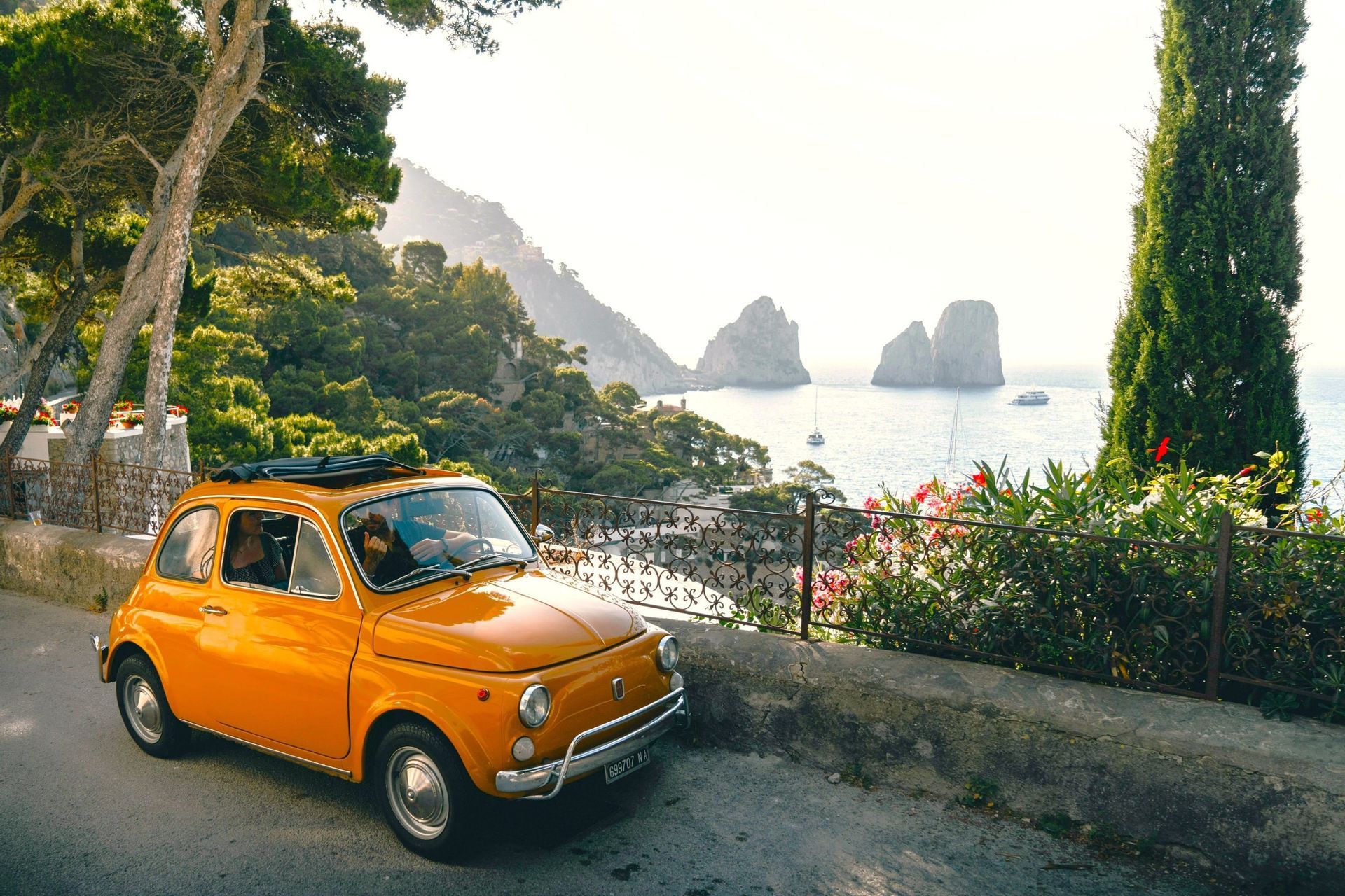An orange vintage car with an open sunroof parked on a scenic coastal road overlooking a sunlit sea with large rock formations.