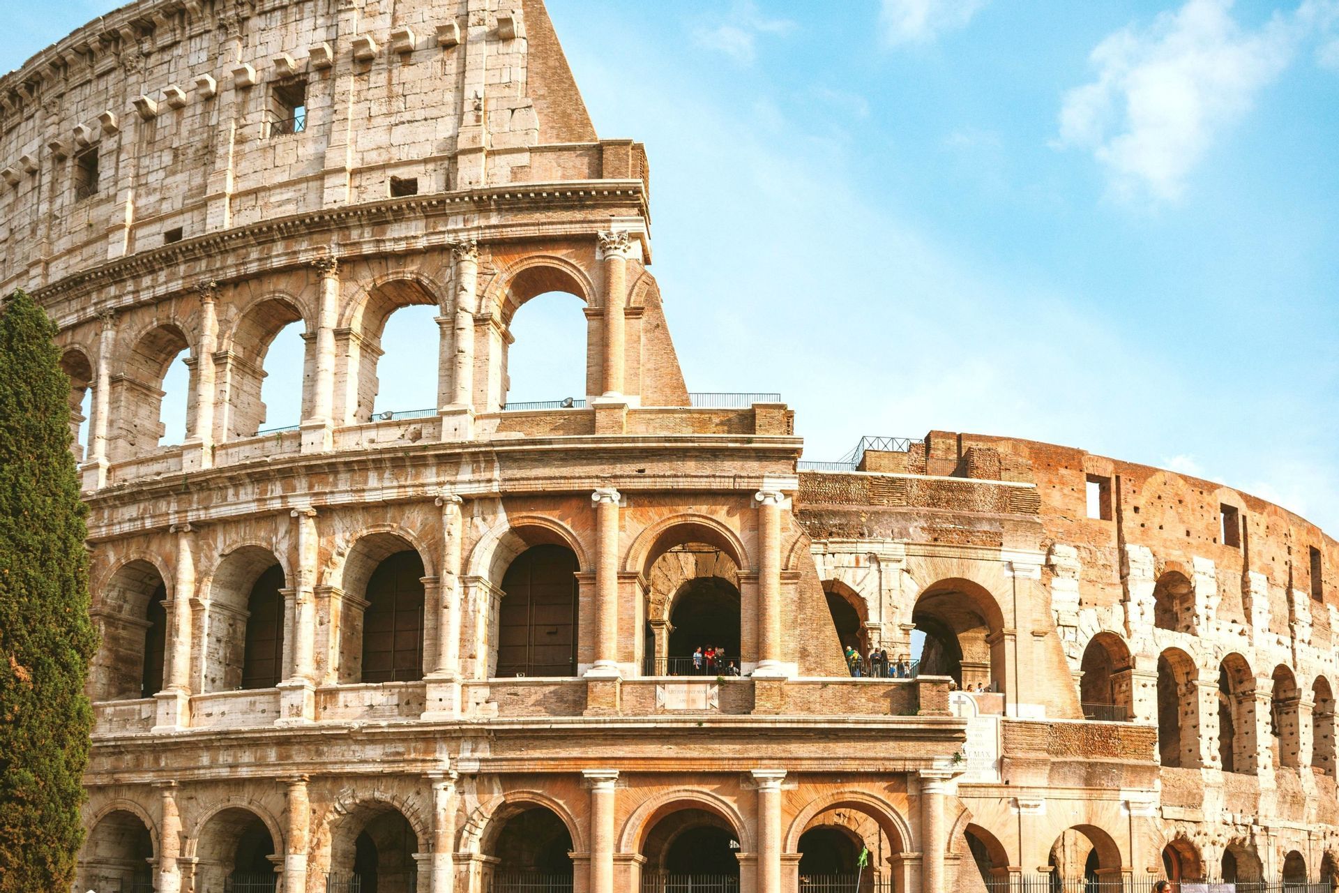 A sunlit, partial view of the ancient Roman Colosseum's tiered stone arches against a bright blue sky with light clouds.