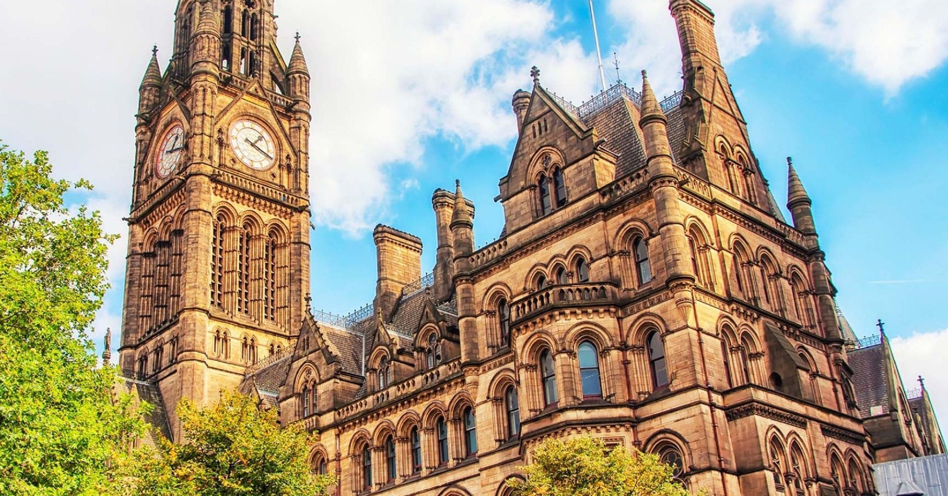 Una vista dal basso di un elaborato edificio in pietra in stile gotico con una grande torre dell'orologio, contro un cielo azzurro con nuvole.