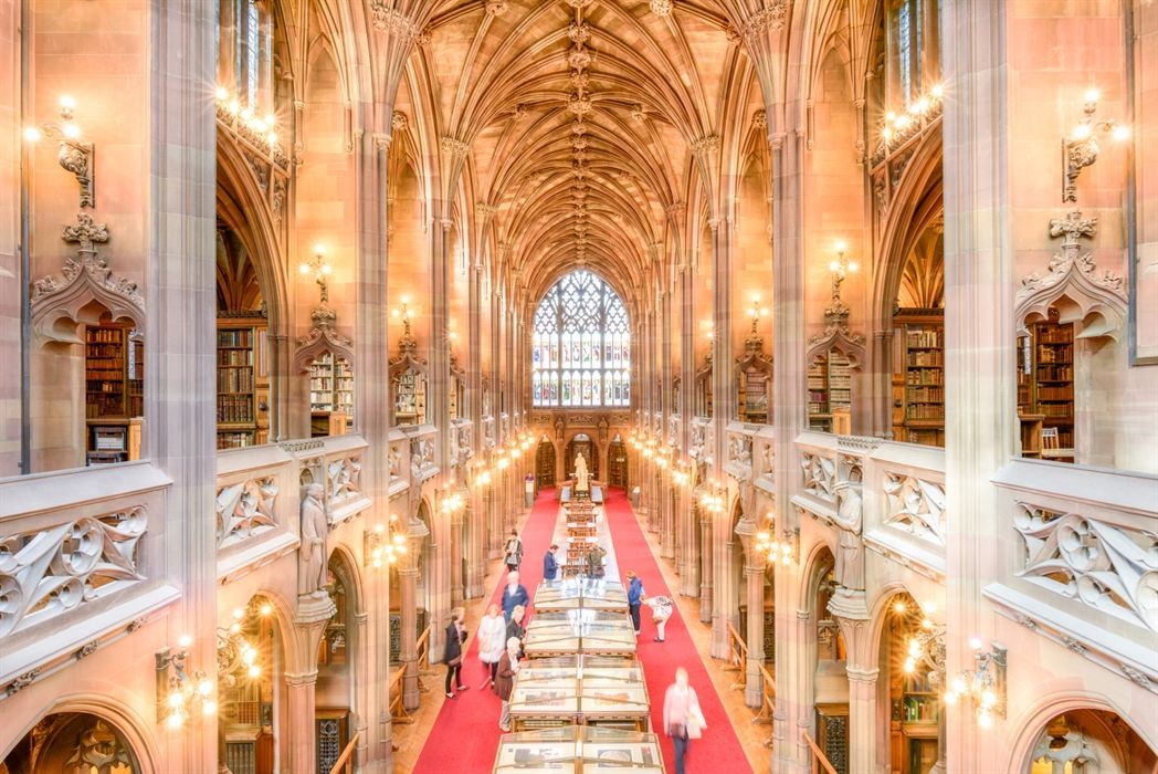 Vista dall'alto di persone che percorrono un tappeto rosso in una magnifica biblioteca storica con alti soffitti a volta e ricca architettura in pietra.