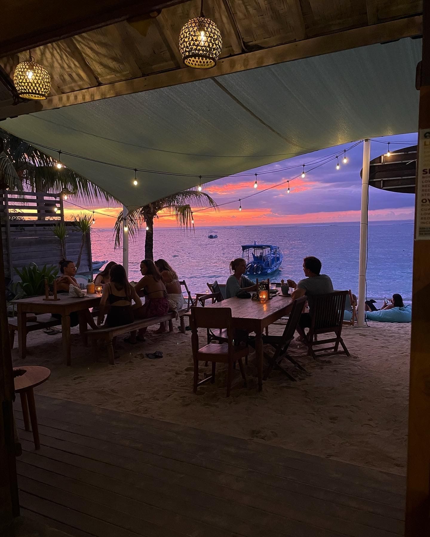 Un gruppo WeRoad cena in un ristorante sulla spiaggia sabbiosa, sotto le lucine, ammirando un vivace tramonto sull'oceano.