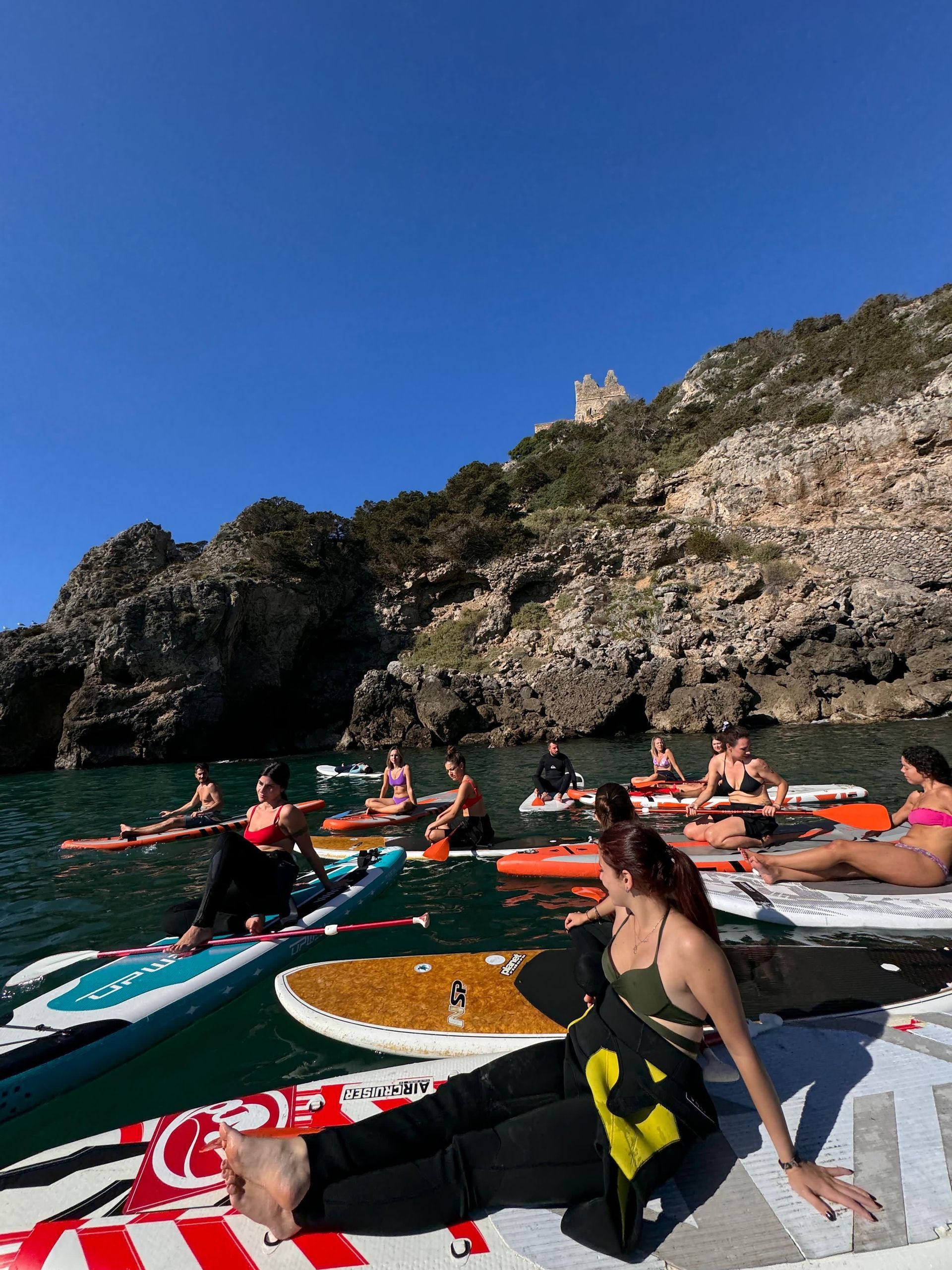 Un viaggio di gruppo WeRoad si rilassa su stand-up paddleboard nel mare, accanto a una scogliera rocciosa con rovine in cima.