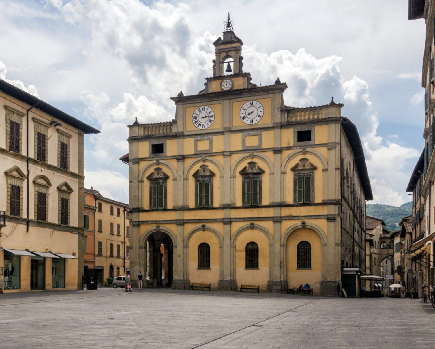Un edificio comunale giallo e ornato con una torre dell'orologio si erge in una grande piazza cittadina pavimentata sotto un cielo nuvoloso.