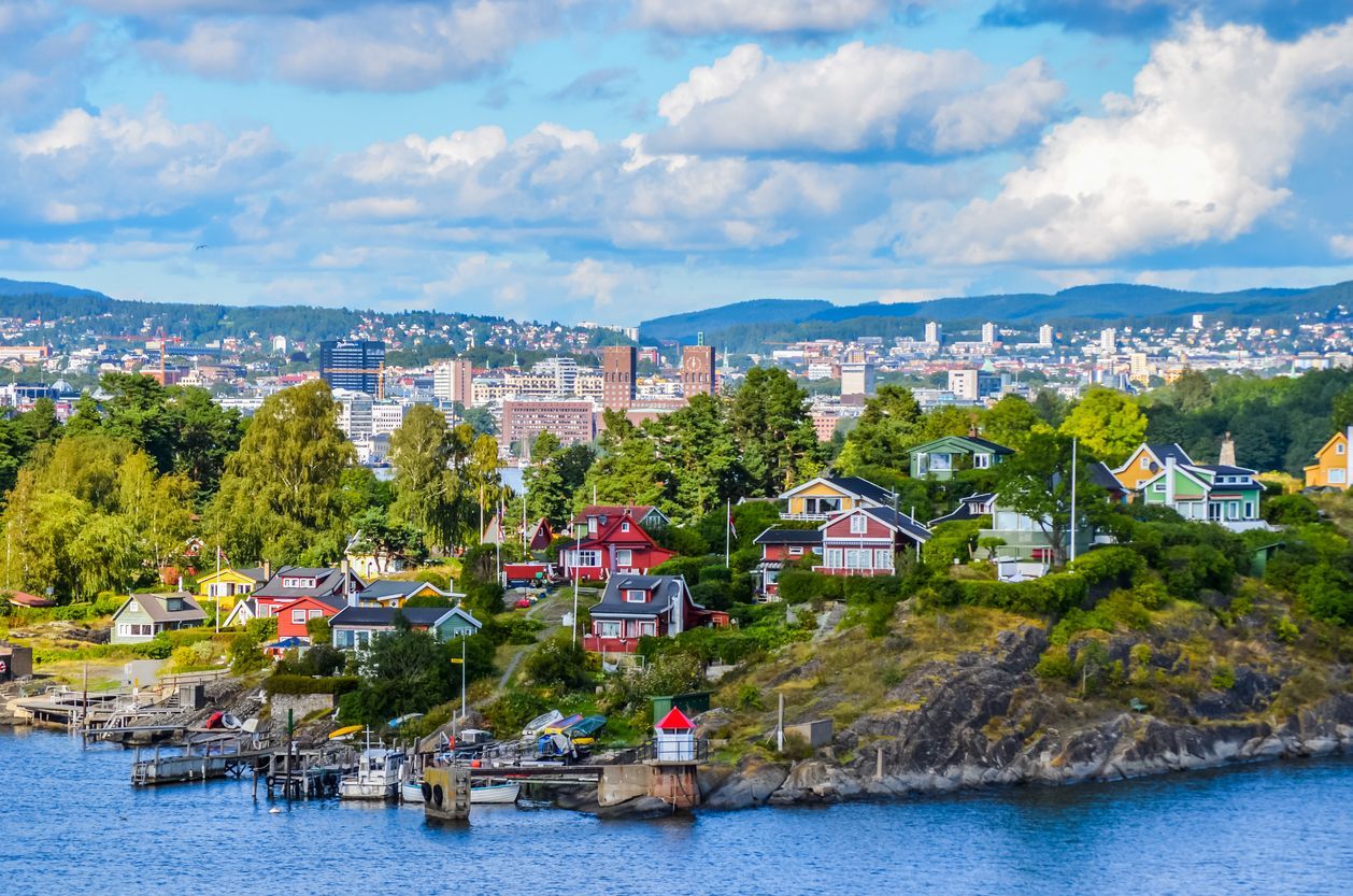 Colorful houses on a rocky, tree-covered coastline with a city in the background under a blue sky with white clouds.