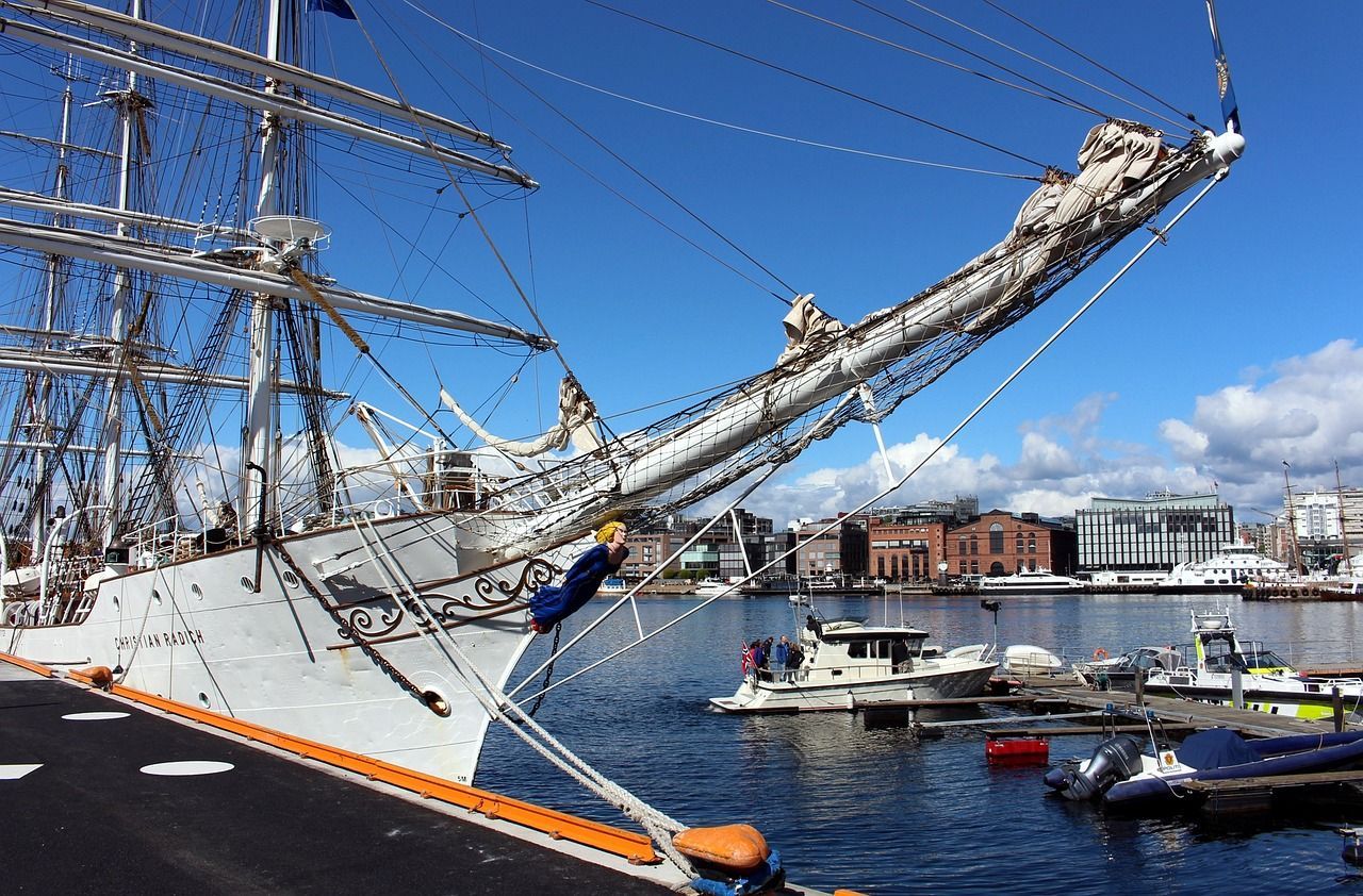 The bow of a large white tall ship with intricate rigging docked in a city harbor under a bright blue sky.