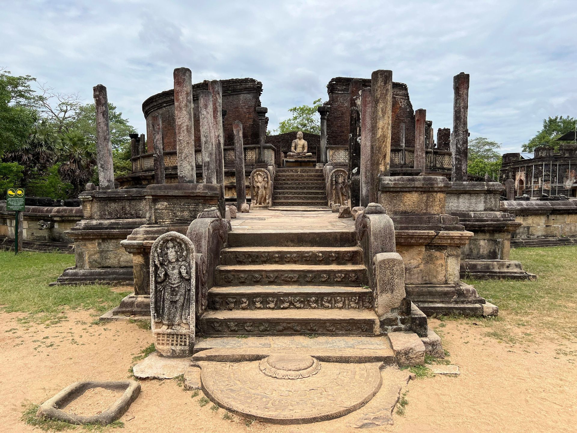 Des marches en pierre mènent à une statue de Bouddha assis au milieu des ruines d'un ancien temple avec de grands piliers en pierre.