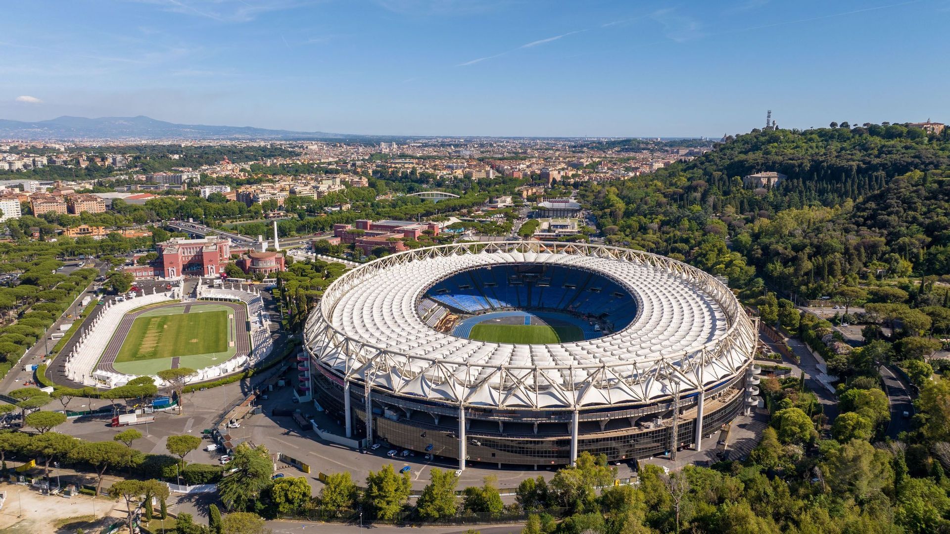 Una vista aerea di un grande stadio sportivo circolare con un tetto bianco, situato accanto a una pista di atletica più piccola ai margini di una città.