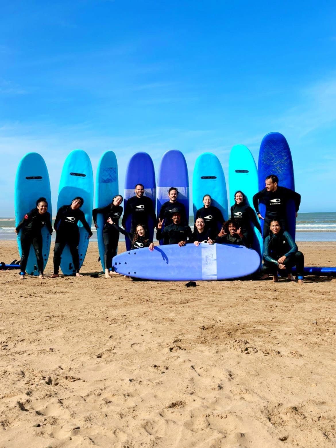 Un gruppo WeRoad in muta posa con le tavole da surf su una spiaggia sabbiosa sotto un cielo azzurro.