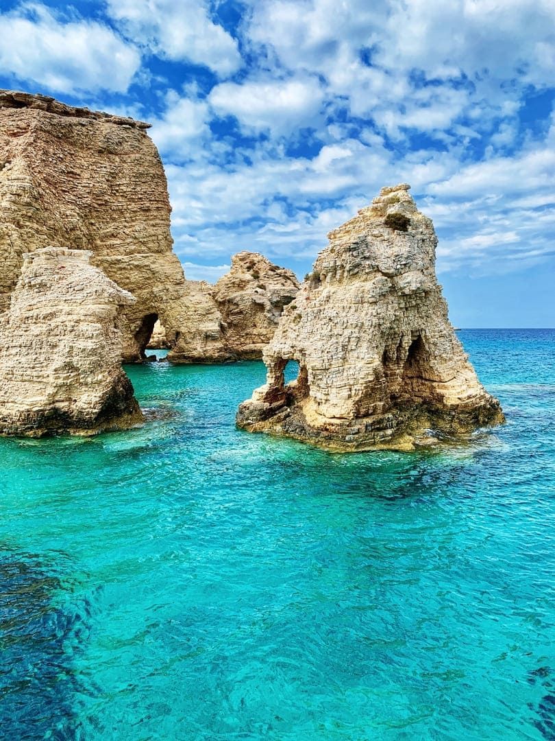 De grandes formations rocheuses avec des arches naturelles émergent d'une eau de mer turquoise et claire sous un ciel bleu partiellement nuageux.