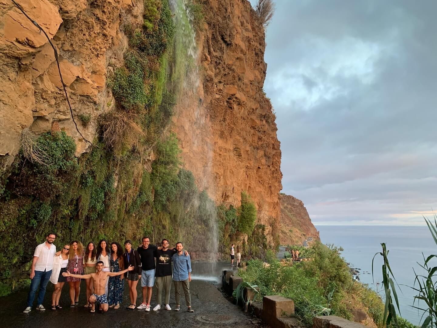 Un gruppo WeRoad in posa su una strada sotto una cascata che scende da una scogliera, con il mare sullo sfondo.