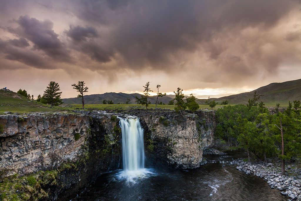 Una cascata si getta da una scogliera rocciosa in un fiume scuro, con verdi pianure e montagne distanti sotto un cielo drammatico e nuvoloso al tramonto.