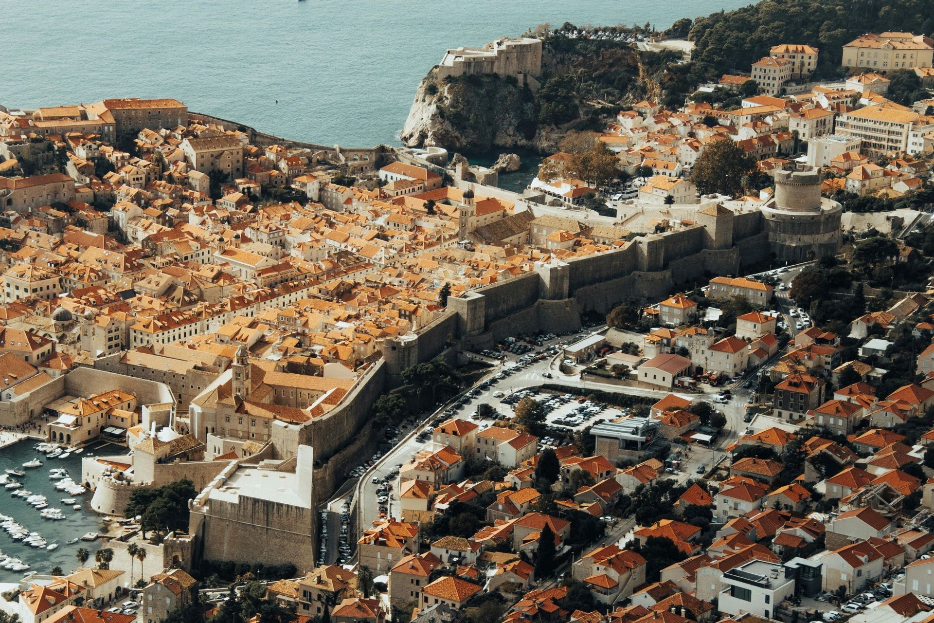 Veduta aerea di una storica città murata con tetti in tegole arancioni, fortificazioni e un piccolo porto sulla costa.