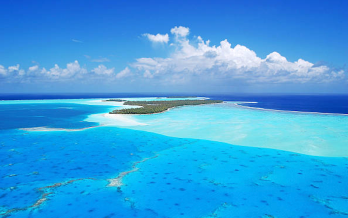 Veduta aerea di un atollo tropicale circondato da una laguna turchese poco profonda e dall'oceano blu intenso, sotto un cielo azzurro con nuvole bianche.