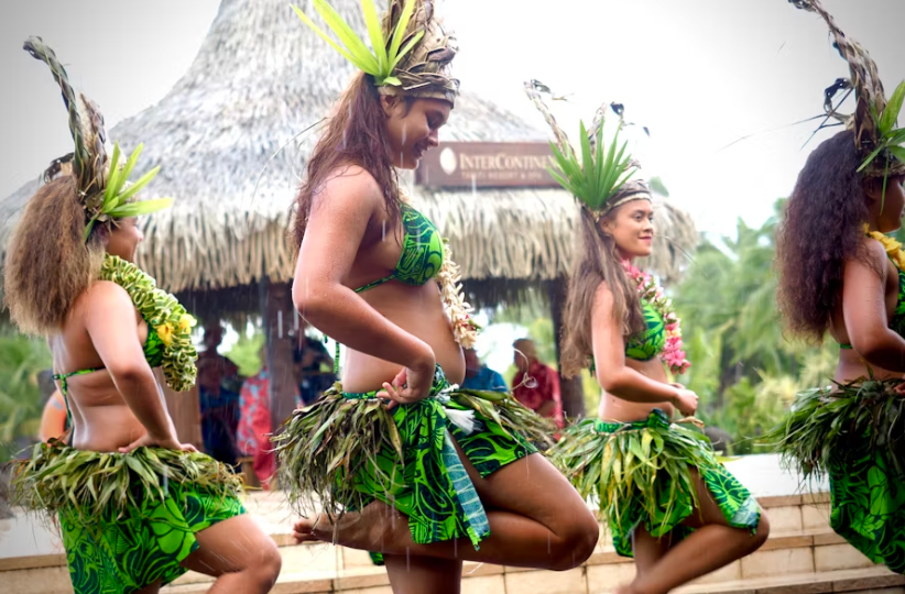 Un gruppo di donne in gonne di foglie verdi e copricapi elaborati esegue una danza tradizionale all'aperto sotto la pioggia.
