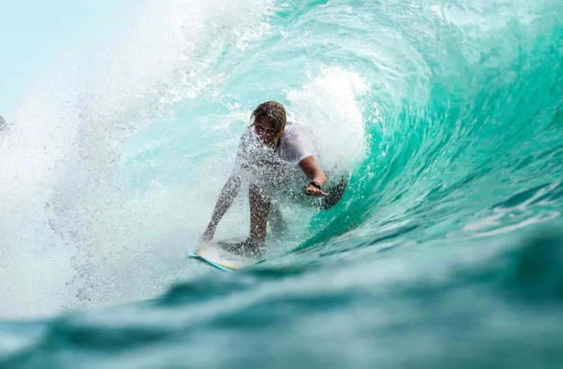 Un surfista con lunghi capelli cavalca la tavola da surf all'interno del tubo di una grande onda turchese che si arriccia.