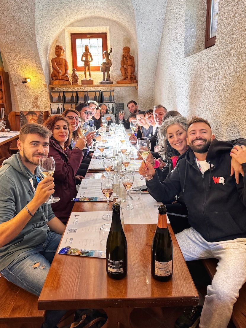 Un viaggio di gruppo WeRoad si gode una degustazione di vini, alzando i calici per un brindisi a un lungo tavolo di legno all'interno di una cantina rustica.
