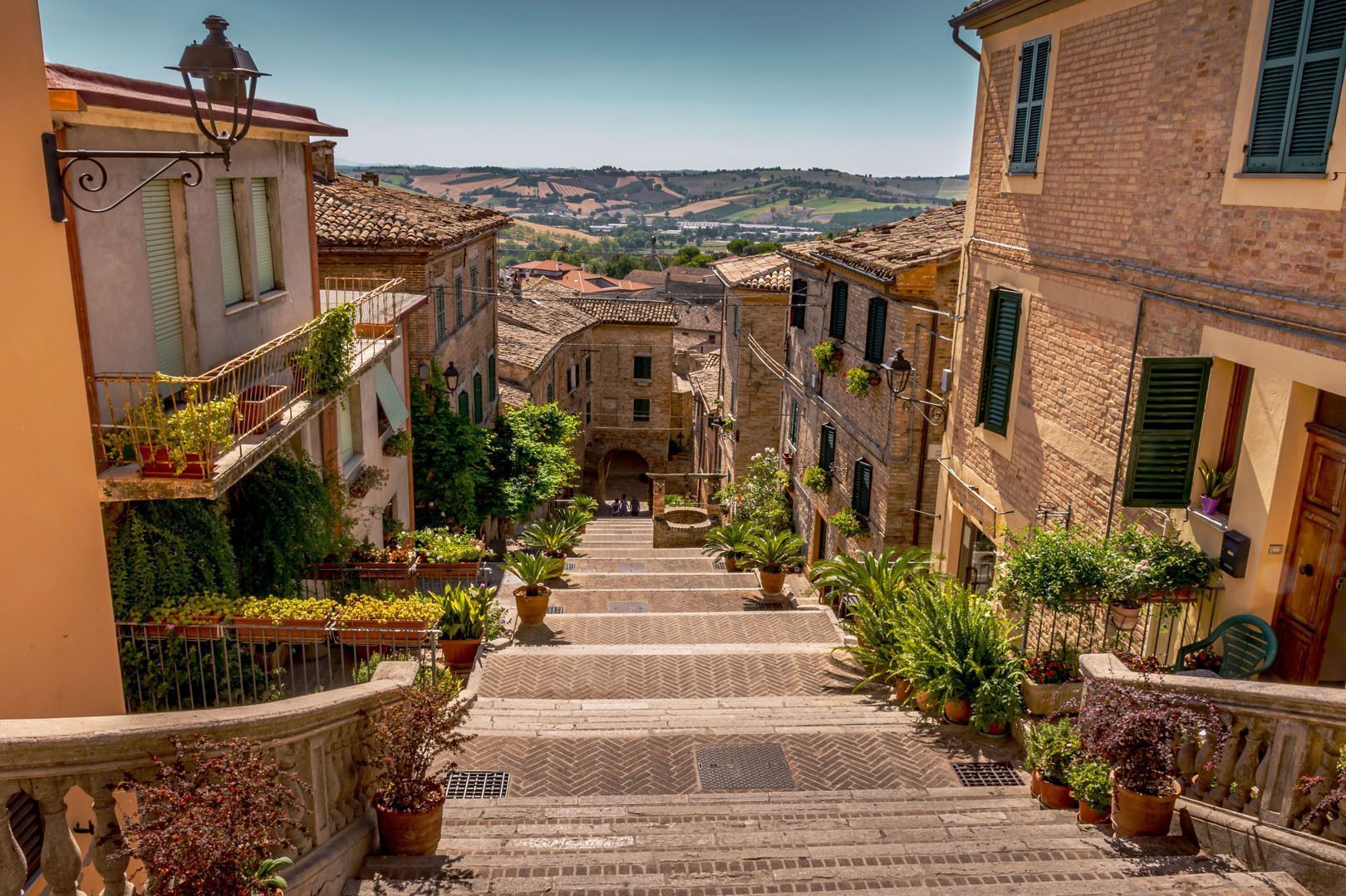 Un'ampia scala in pietra scende lungo una stretta via del paese, fiancheggiata da piante in vaso ed edifici in mattoni, con dolci colline sullo sfondo.