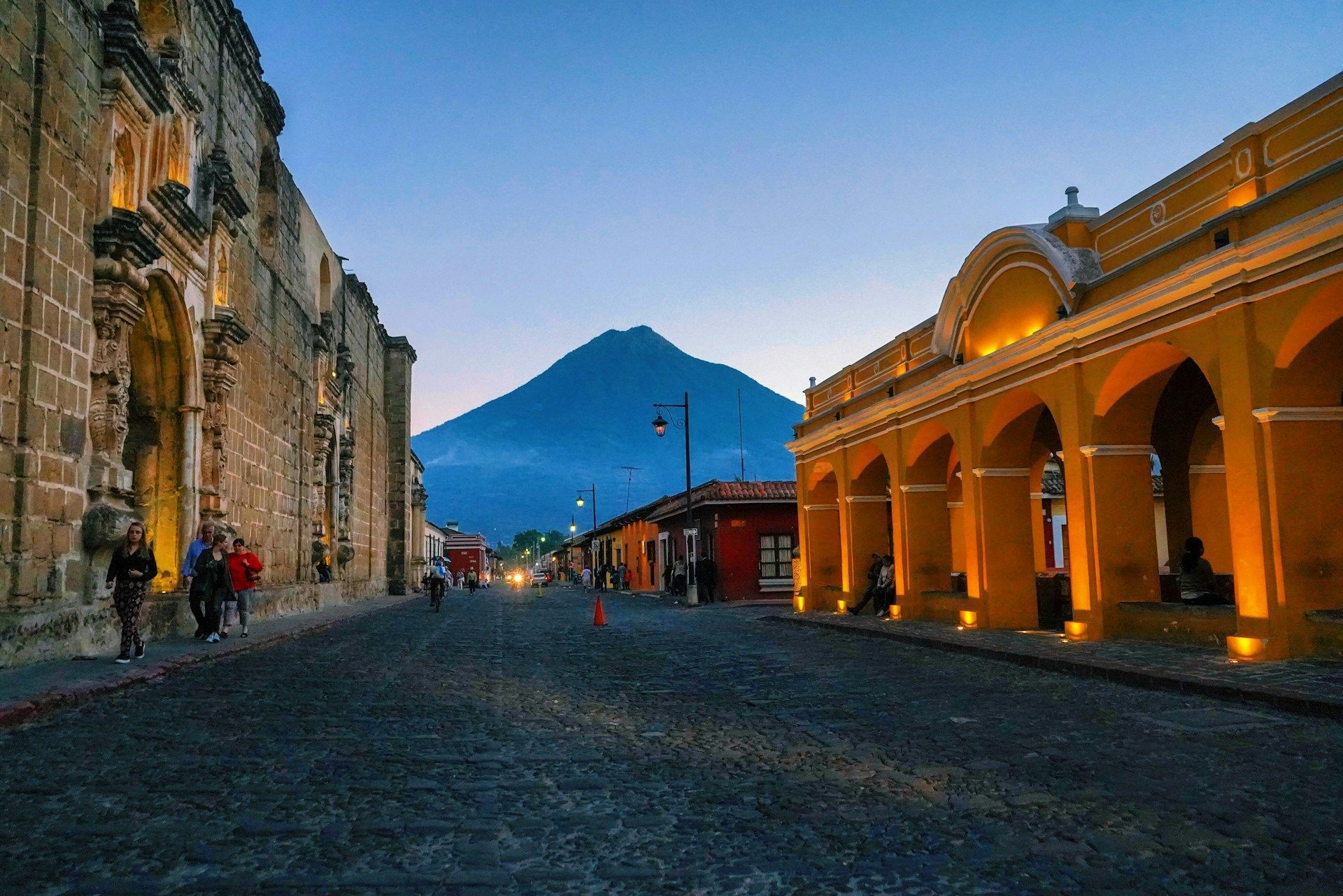 Una calle empedrada flanqueada por edificios coloniales iluminados conduce hacia un gran volcán al atardecer.