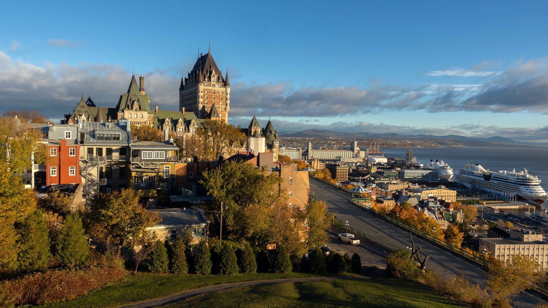 Un paysage urbain avec un bâtiment historique ressemblant à un château, des arbres d'automne et un port fluvial avec des navires de croisière amarrés.