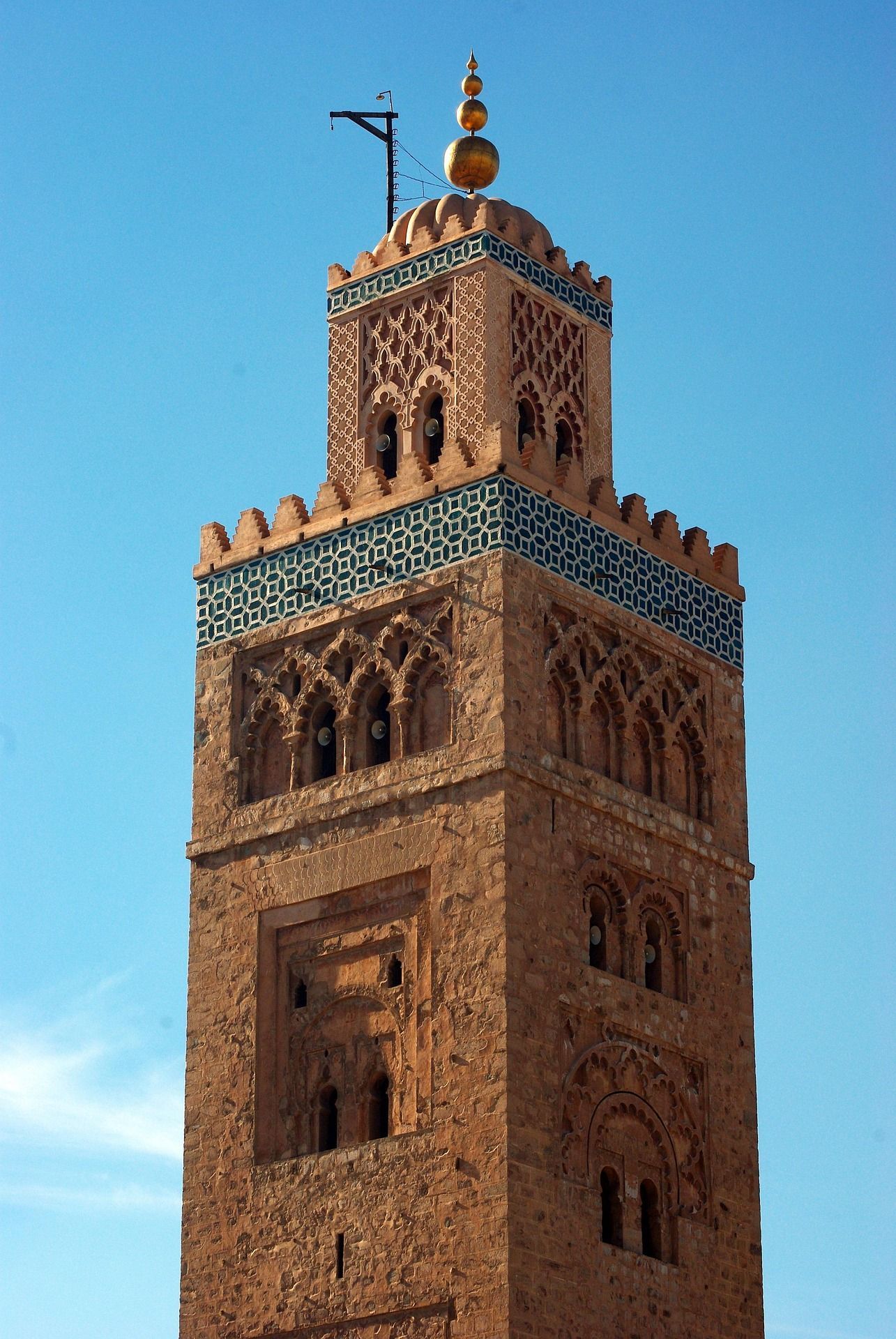 Un minarete ornamentado de piedra rojiza con tallas detalladas y azulejos geométricos azules bajo un cielo azul claro