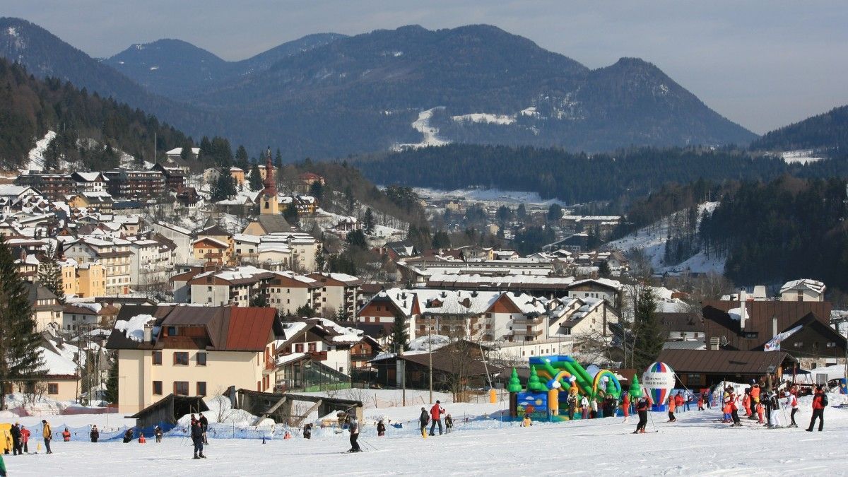 Un'ampia veduta di una località sciistica coperta di neve, con persone che sciano su una pista in primo piano e montagne boscose sullo sfondo.