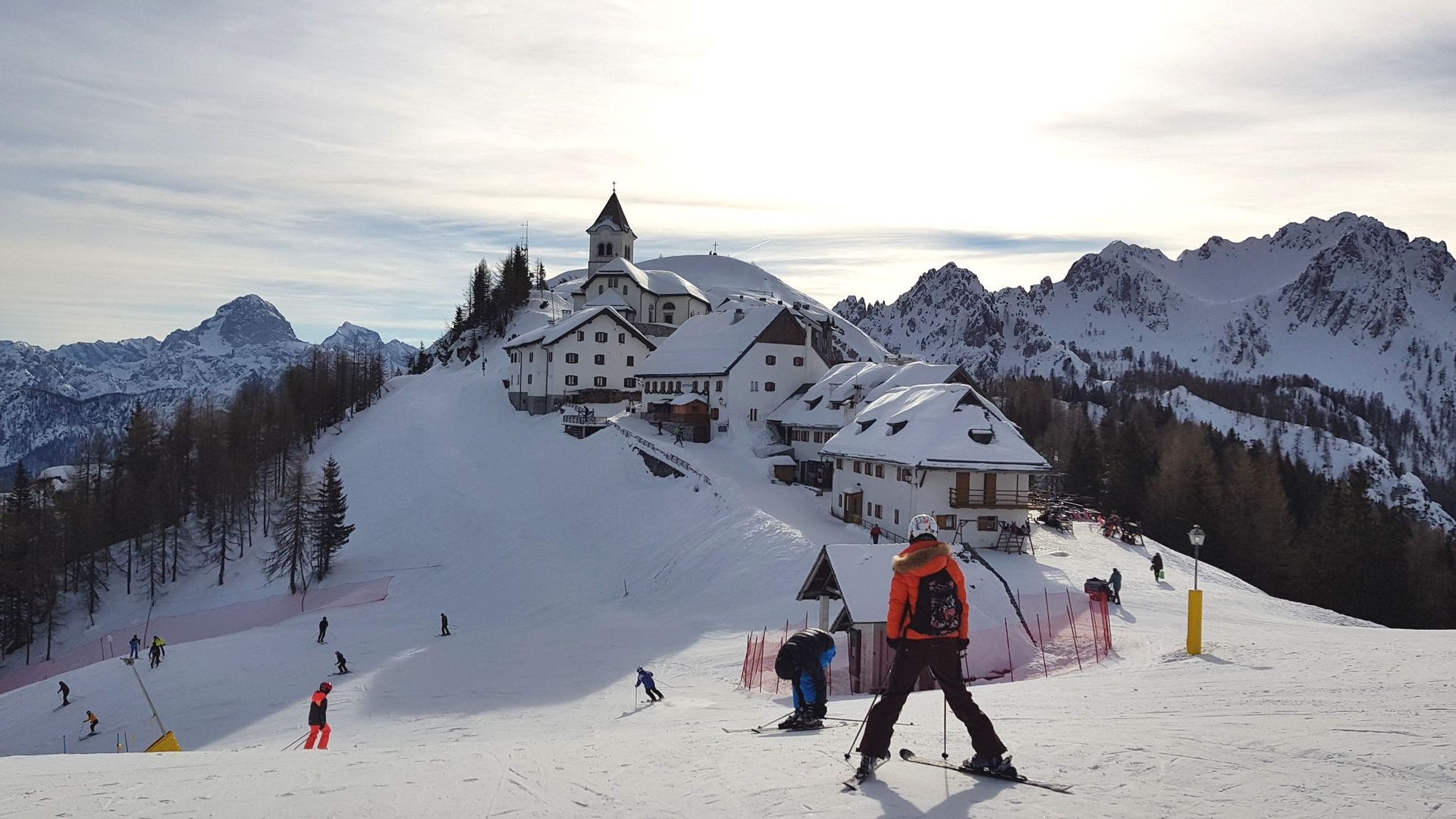 Sciatori su una pista innevata con un villaggio sormontato da una chiesa e cime montuose sullo sfondo sotto un cielo soleggiato.