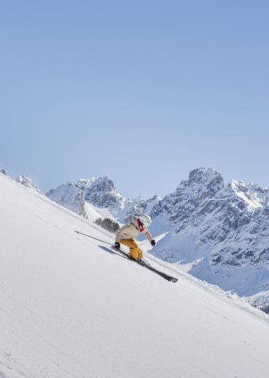 Una persona scia su un'ampia pista innevata, con montagne coperte di neve sullo sfondo e un cielo sereno.
