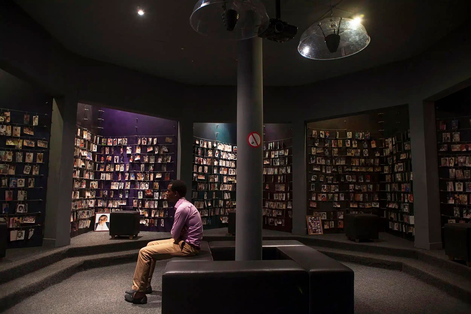 Un homme est assis sur un banc dans une salle commémorative circulaire faiblement éclairée, contemplant des murs ornés de nombreux portraits photographiques.