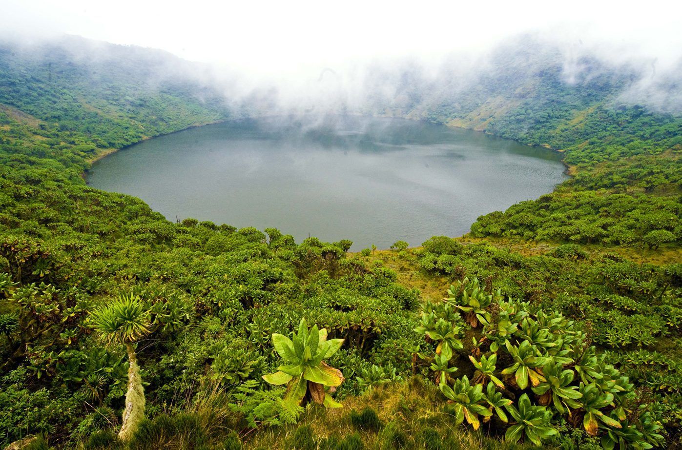 Vue plongeante d'un lac de cratère circulaire, entouré de collines à la végétation luxuriante et verte, sous un ciel brumeux et couvert.