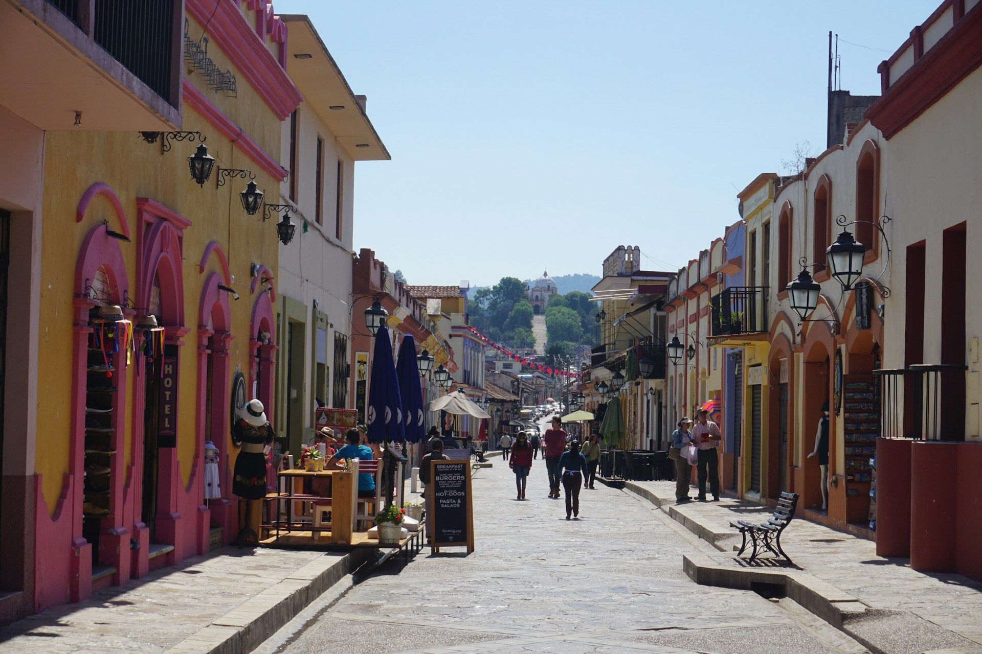 Persone passeggiano lungo una strada acciottolata e assolata, fiancheggiata da colorati edifici coloniali, con vista su una struttura su una collina lontana.