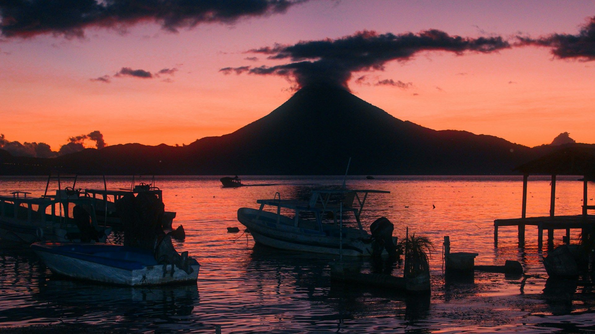Diverse barche galleggiano su un lago al tramonto con una grande montagna in silhouette sullo sfondo sotto un cielo arancione e viola.