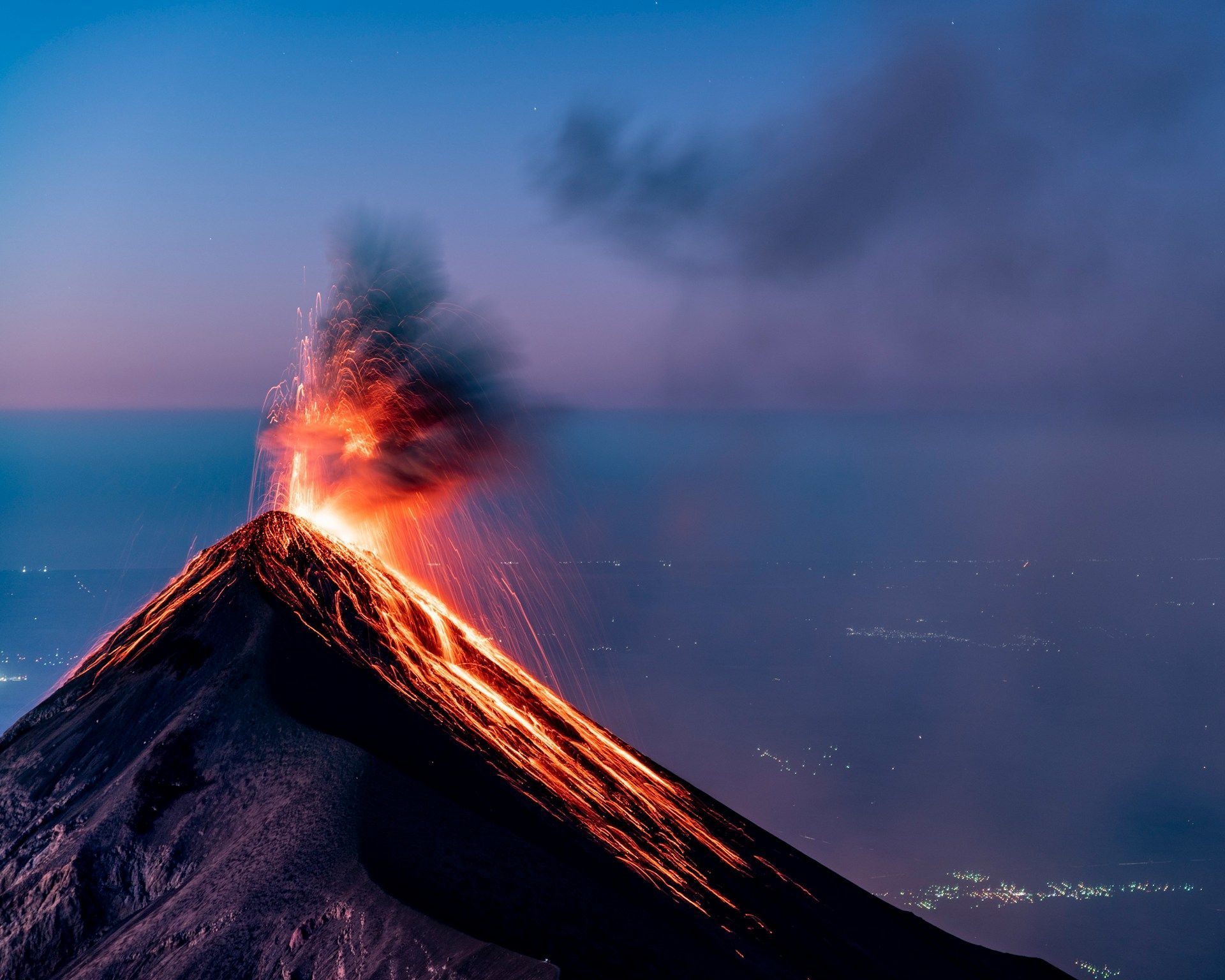 Un vulcano erutta al tramonto, con lava fusa che scorre lungo i suoi pendii e scintille che si alzano nel cielo sopra un paesaggio di luci cittadine.