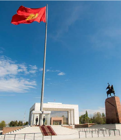 La bandiera del Kirghizistan sventola su un'alta asta in una piazza, con due guardie alla sua base davanti a un edificio moderno e a una statua equestre.