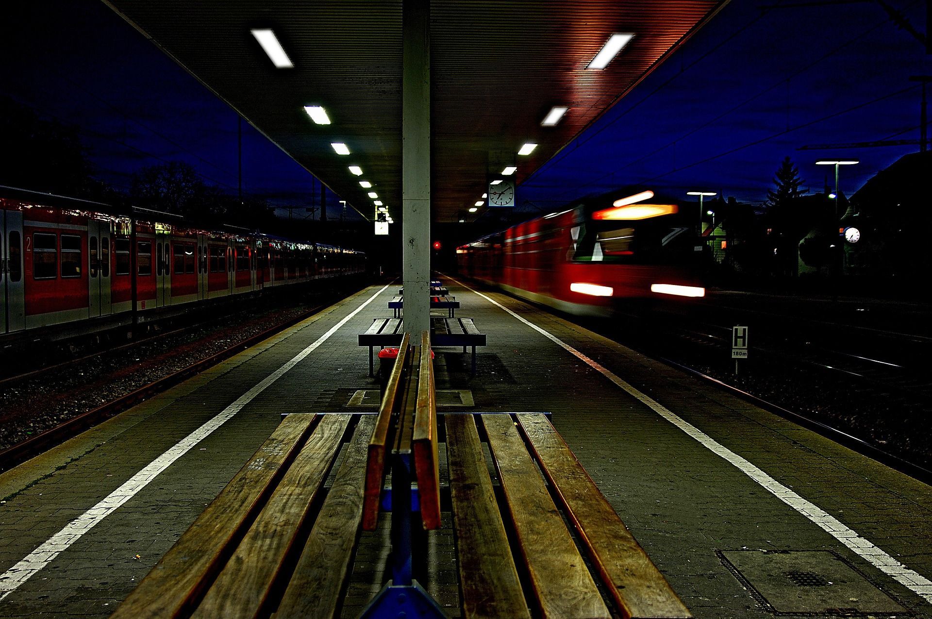 Un treno rosso sfreccia di notte davanti a una banchina della stazione vuota e illuminata, creando un effetto di mosso.