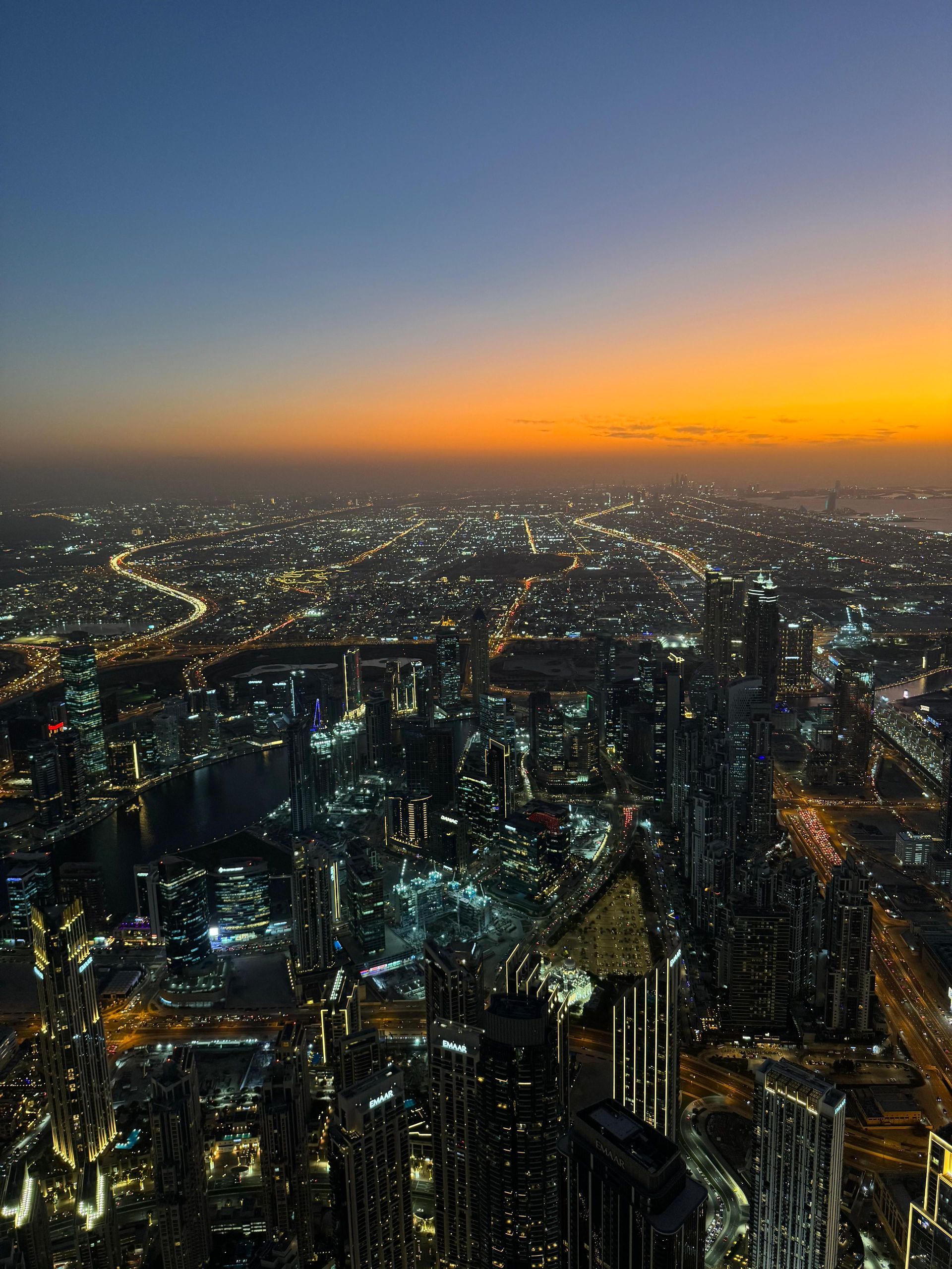 Veduta aerea di una metropoli estesa al crepuscolo, con grattacieli e autostrade illuminati sotto un cielo sfumato arancione e blu.
